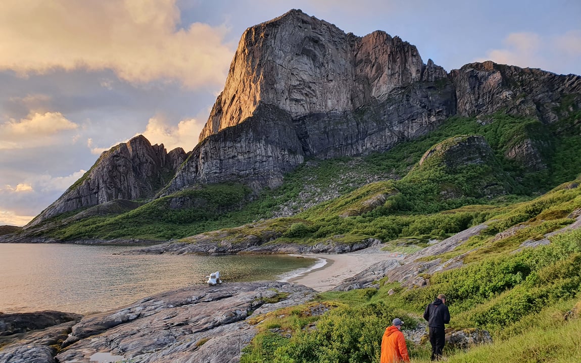 To personer går mot et fjell, med strand til venstre og grønn vegetasjon til høyre i bildet.