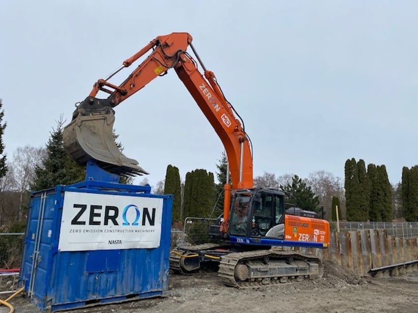 En oransje gravemaskin lener grabben på toppen av en blå kontainer med skriften "ZERON" på siden