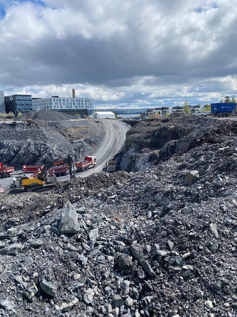 Et stort område med steinmasser i forskjellige størrelser. Nede i gropa står en gravemaskin og to lastebiler med henger. I bakgrunnen står flere bygg.